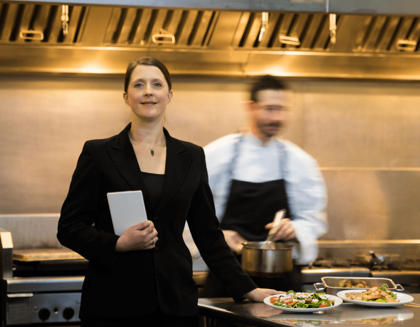 A woman in a suit, holding a notepad, in a restaurant kitchen