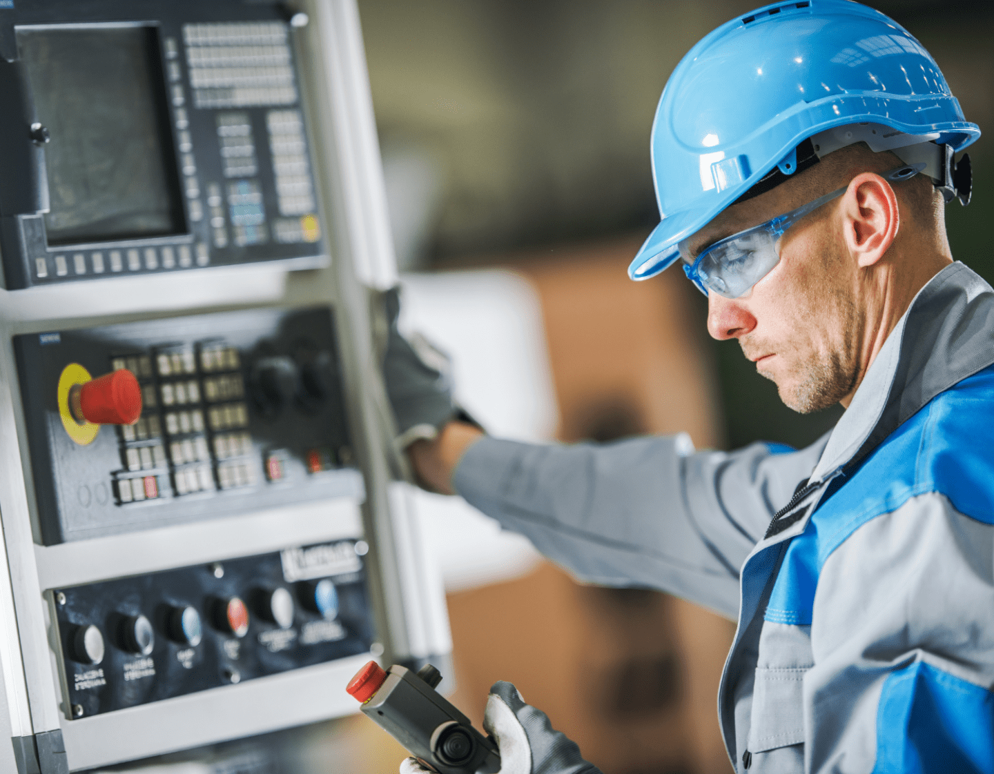 An image of a worker in a factory using a CNC machine. The worker is wearing blue protective gear, including a hard-hat and glasses. He is looking at readings on a hand-held device