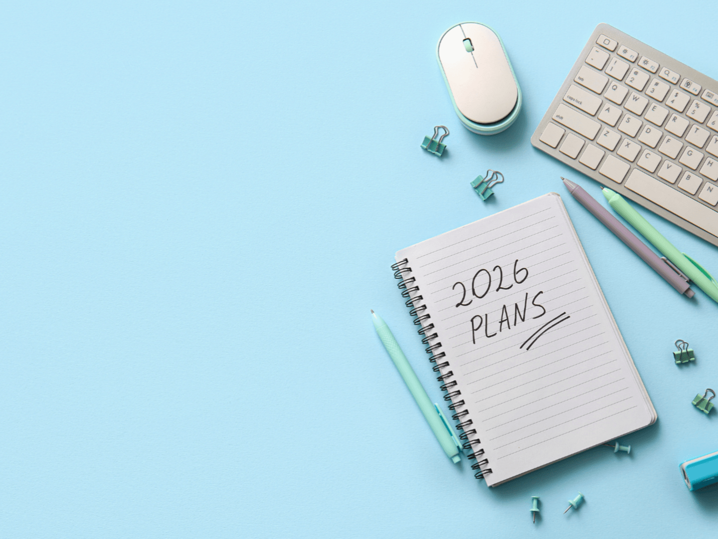 An image of a notebook, pens, a computer mouse and keyboard, and paperclips on a light-blue background. The notebook has the words "2026 Plans" written on it.