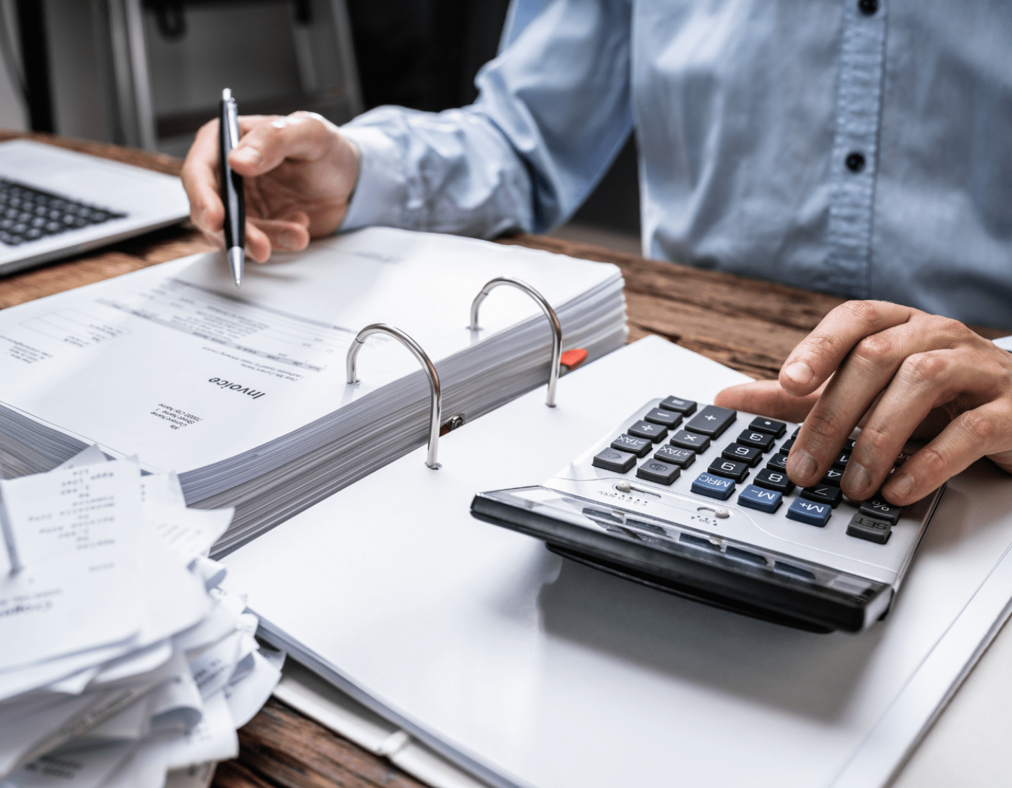 A close-up image of a man working on accounts. He is sat at a wooden desk, and is using a calculator while reading through a folder of invoices.