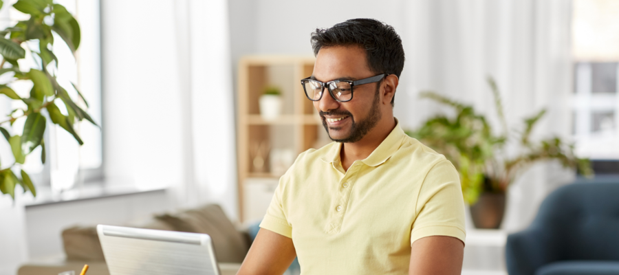 An image of a man sitting at a desk and using a laptop. He is wearing black-framed glasses and a yellow polo shirt