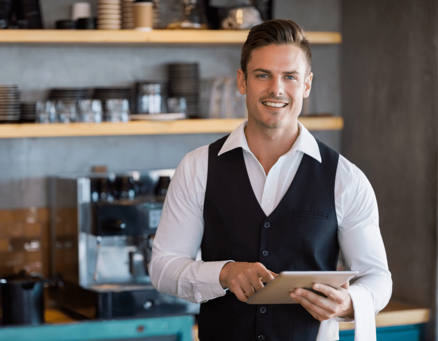 An image of a front of house worker using an ipad. He is dressed smartly and is standing near a coffee making machine