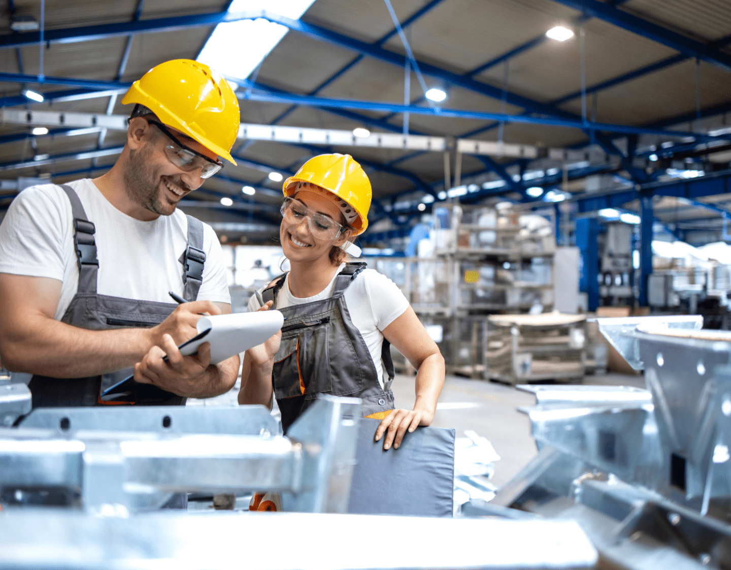 An image of a Production Manager speaking to a worker in a factory. The manager is holding a clipboard and they are standing next to large metal components.