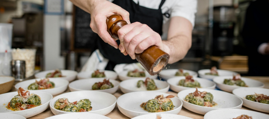 A close up image of a chef seasoning a lot of small dishes. The chef is wearing chef whites and is using a wooden pepper mill.