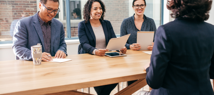 An image of 3 people interviewing one person for a job. The interviewers are sat on one side of a wooden table, and have the person's CV in their hands.