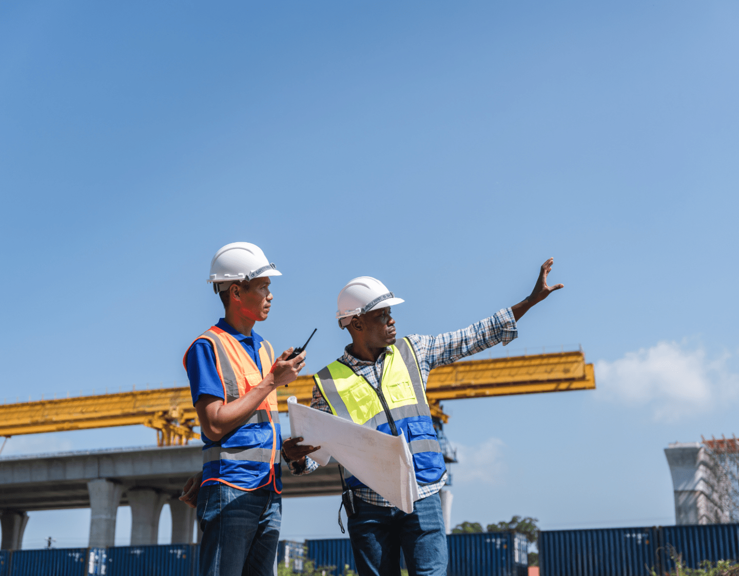 An image of two workers discussing a large-scale engineering project outside in a city. They are wearing high-visibility vests and hard-hats.