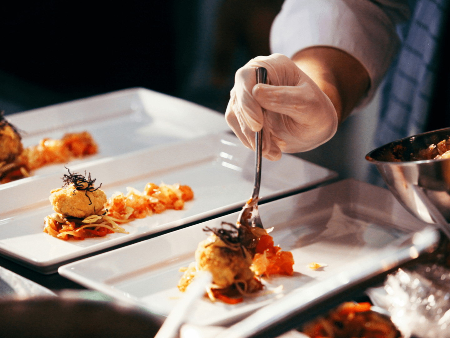 A close up image of a chef plating several dishes at once. They are holding a spoon and frying pan, and placing ingredients on to the plates with a spoon