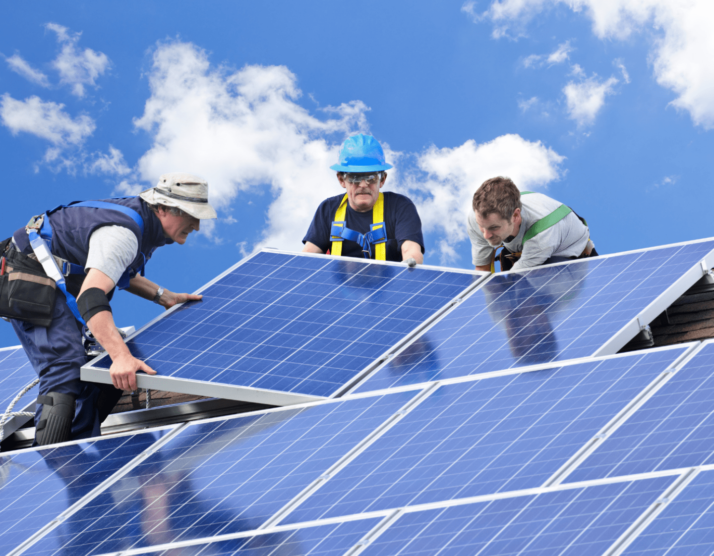An image of three workers installing solar panels on the roof of a house. They are wearing hard hats and harnesses with tools on their belts.