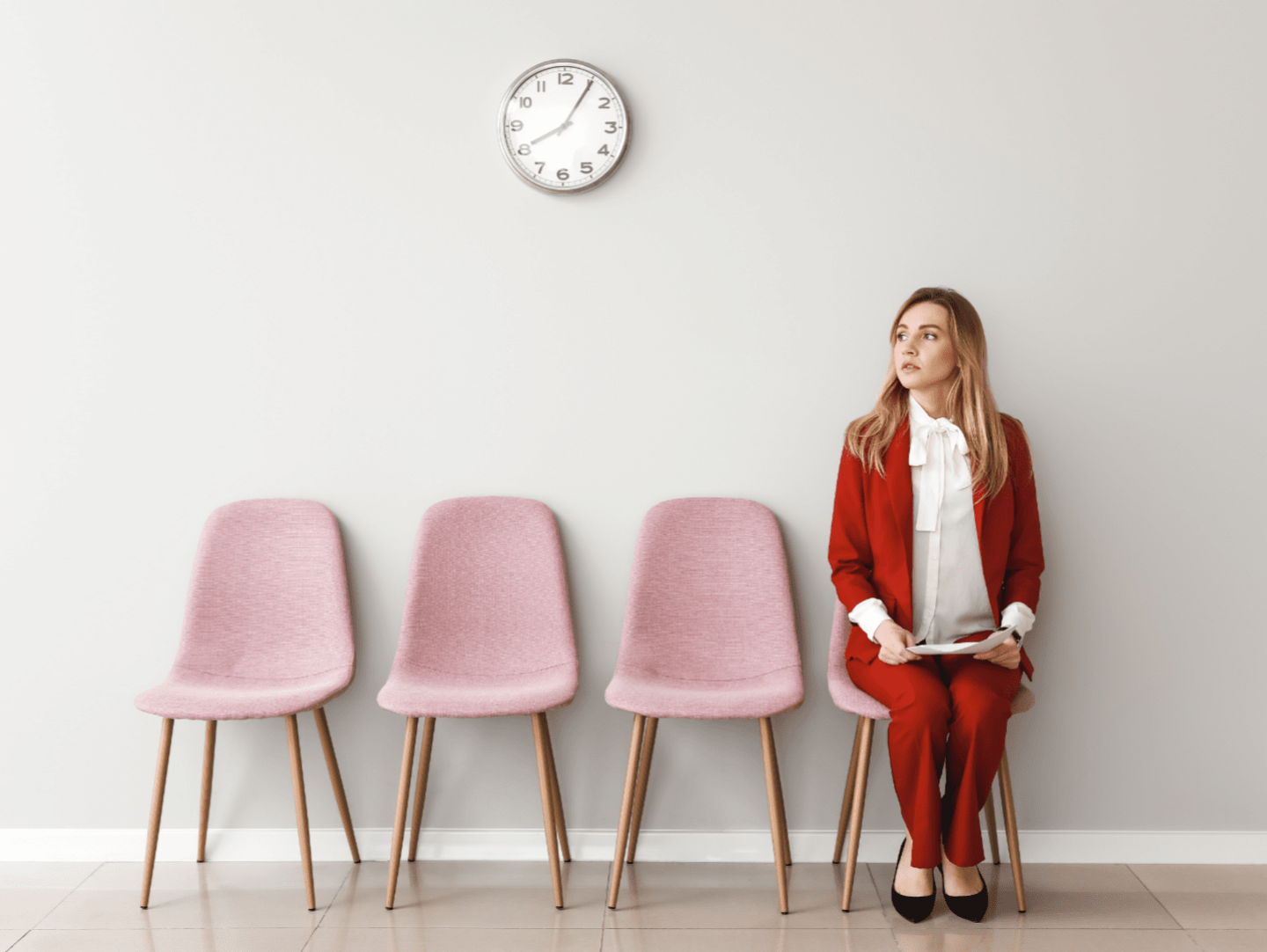 An image of a woman waiting for a job interview. She is wearing a red suit with a white shirt and is sitting on a pink chair. She has her CV in her hand and is looking towards the interview room. There are 3 empty chairs to her right.