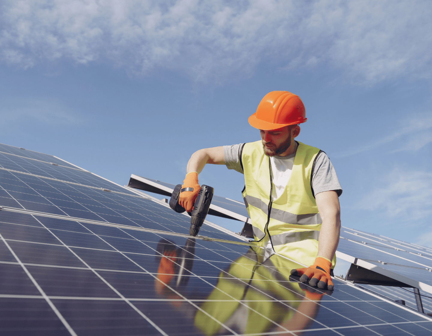 An image of a solar panel installer working on a roof. He is screwing panels in to place, and wearing high-visibility construction wear.